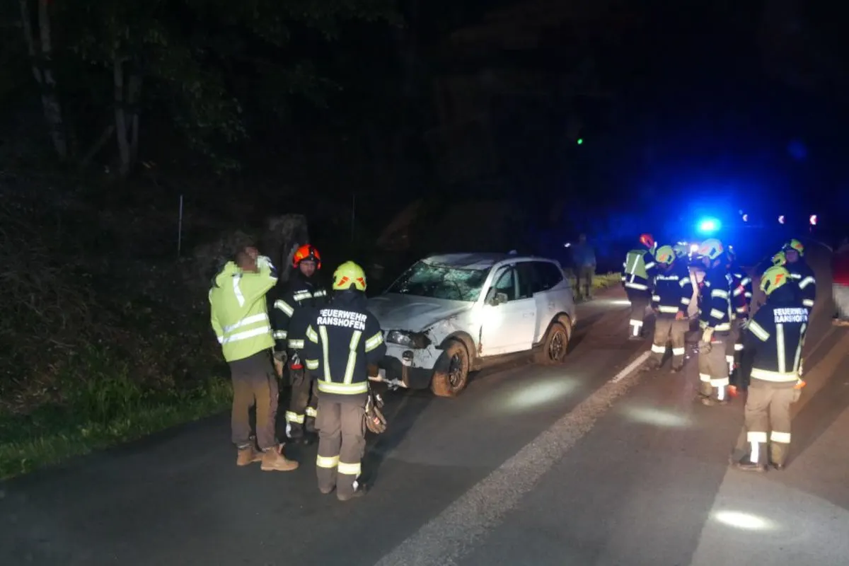 Ein weißes Auto mit sichtbaren Schäden steht auf einer Straße, umgeben von Feuerwehrleuten mit Schutzhelmen in der Nacht.