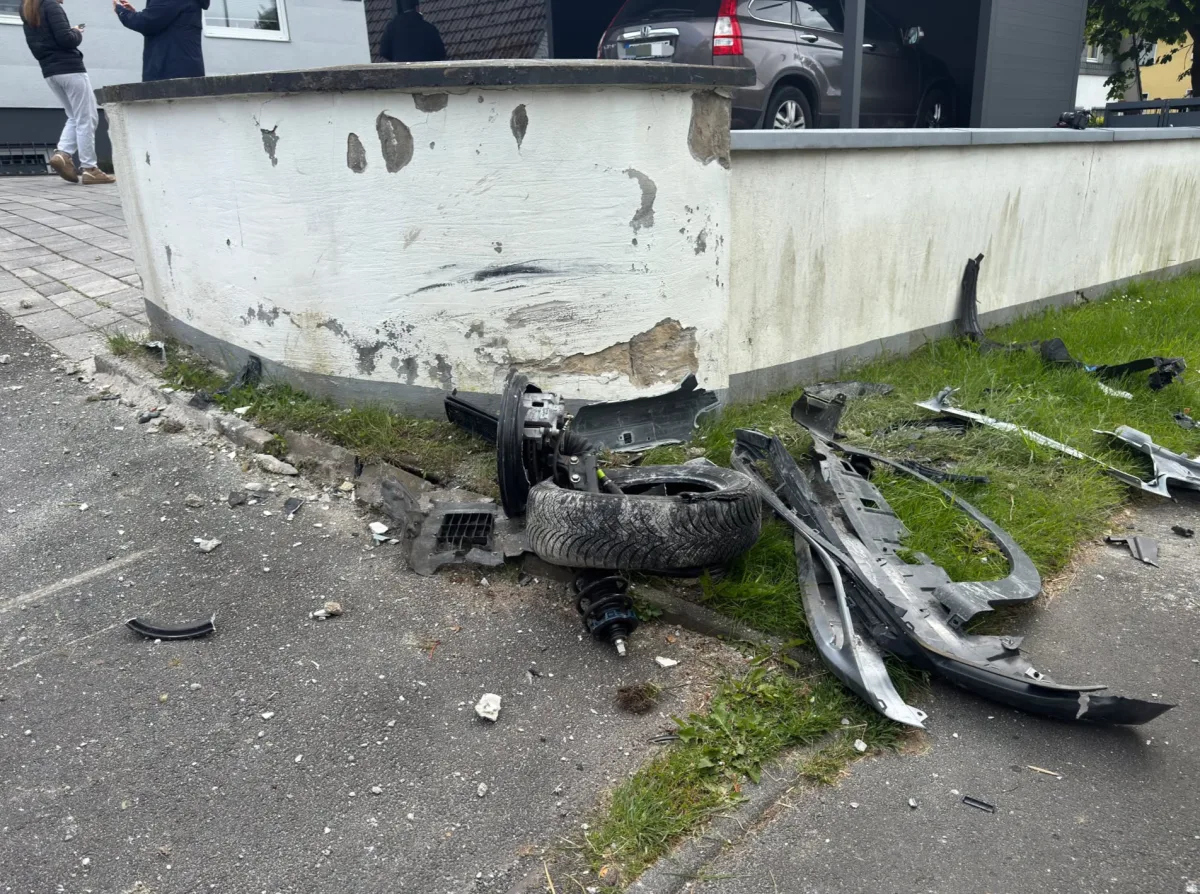 Eine beschädigte Mauer in einer Wohngegend, daneben liegen mehrere Autoteile verstreut auf dem Boden. Wenige Menschen stehen im Hintergrund.