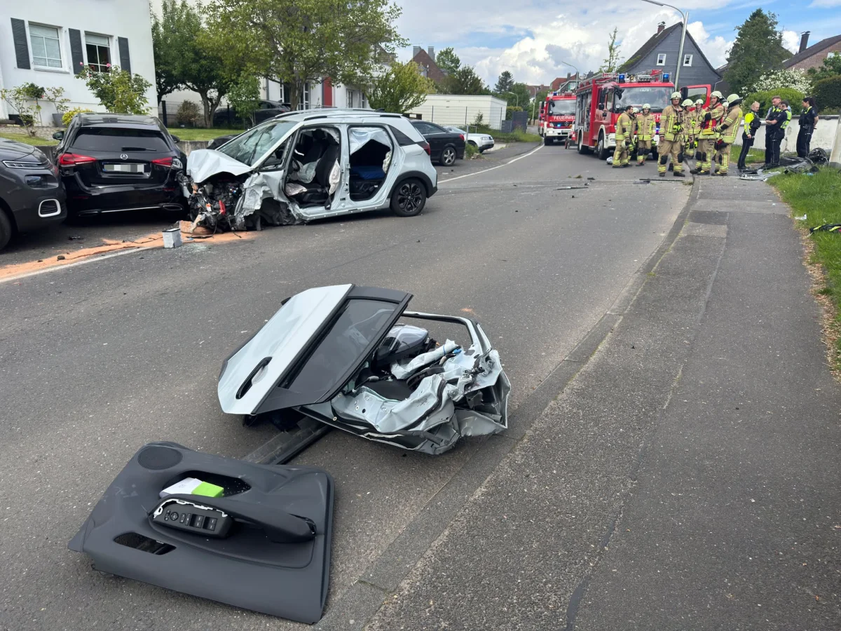 Zerstörte Fahrzeugtüren liegen vor zwei stark beschädigten Autos. Feuerwehrleute stehen in einer Wohnstraße bei einem Einsatzfahrzeug der Feuerwehr.