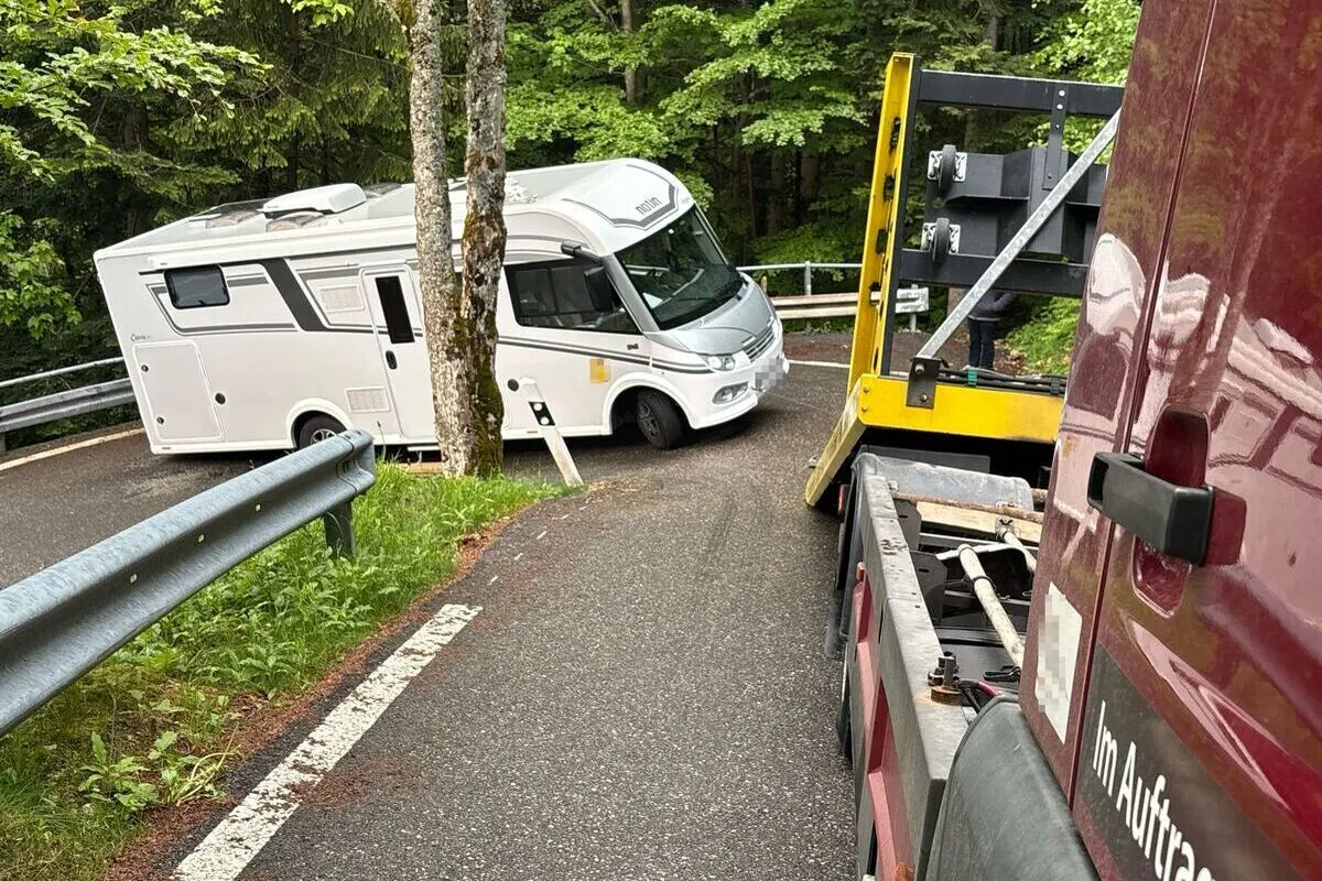 Ein weißes Wohnmobil steht quer über eine schmale Straße im Wald und wird von einem Abschleppfahrzeug hinter einem Baum geborgen.