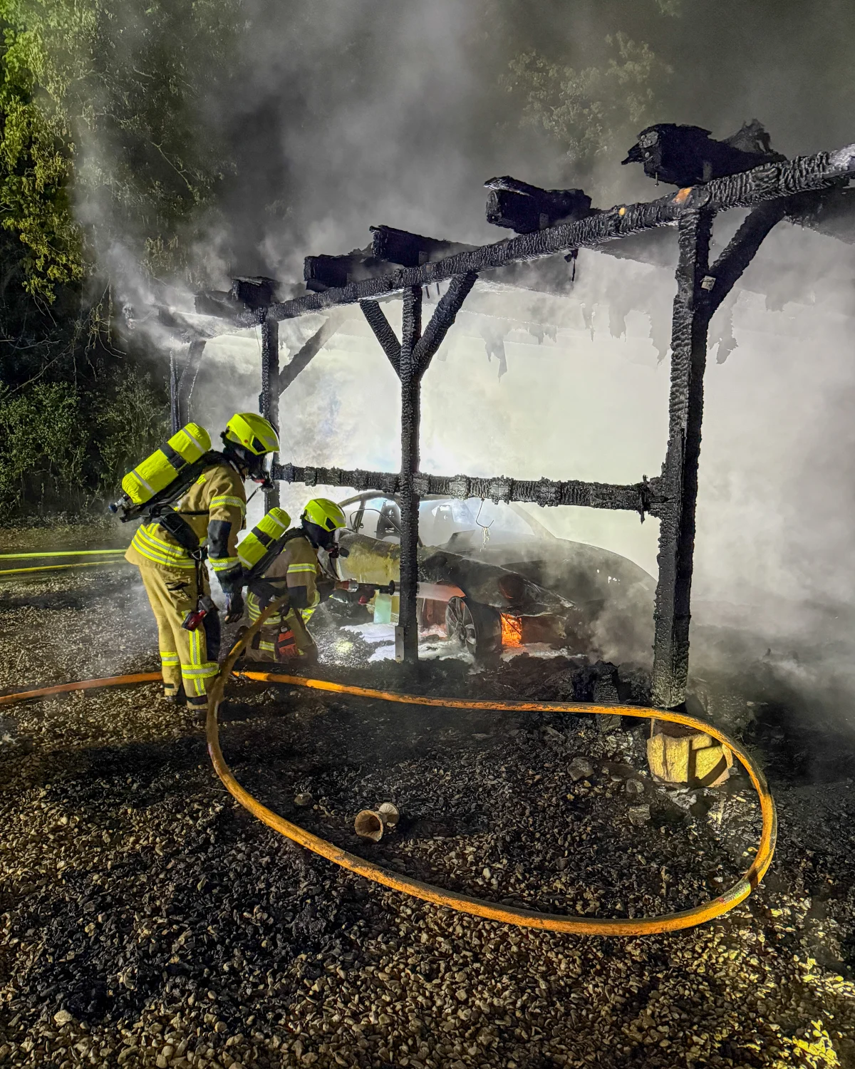 Zwei Feuerwehrleute mit Atemschutzgeräten löschen bei Nacht ein brennendes Auto unter einem stark beschädigten Carport. Rauch und Flammen sind sichtbar.