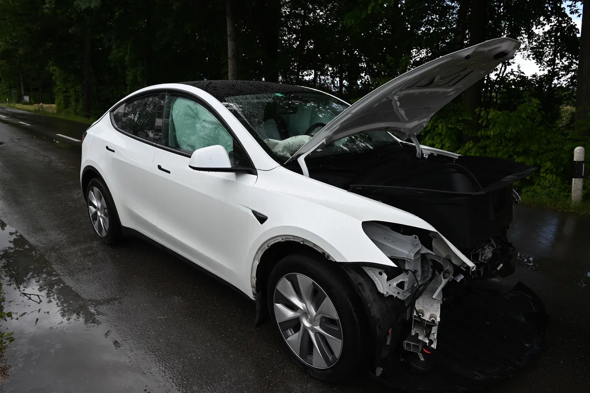 Ein weißes Auto mit offenem Motorraum und Frontschaden parkt am regennassen Straßenrand in einer bewaldeten Umgebung.