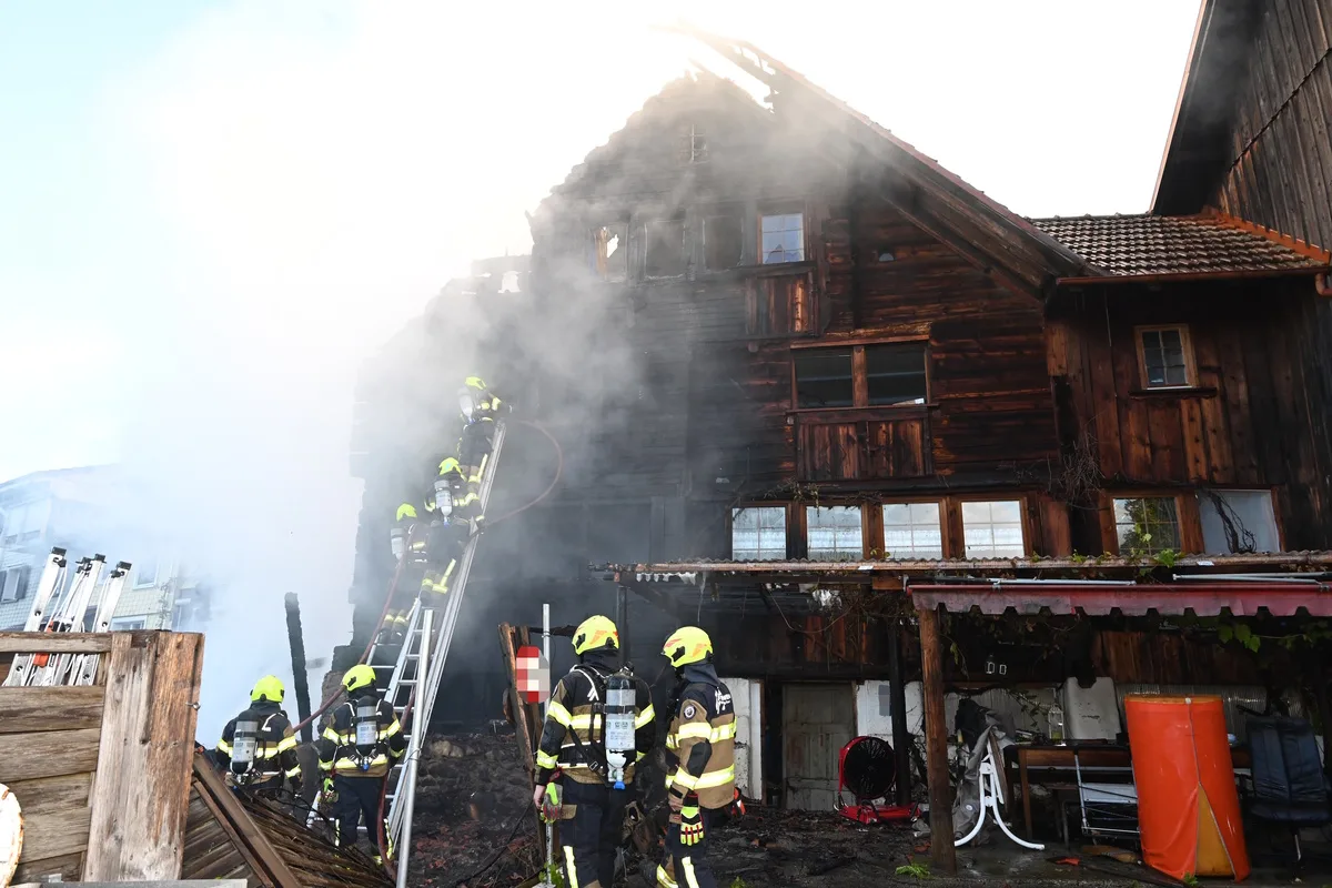 Mehrere Feuerwehrleute in Ausrüstung bekämpfen einen Brand in einem alten, mehrstöckigen Holzhaus mit Leitern und Schläuchen.