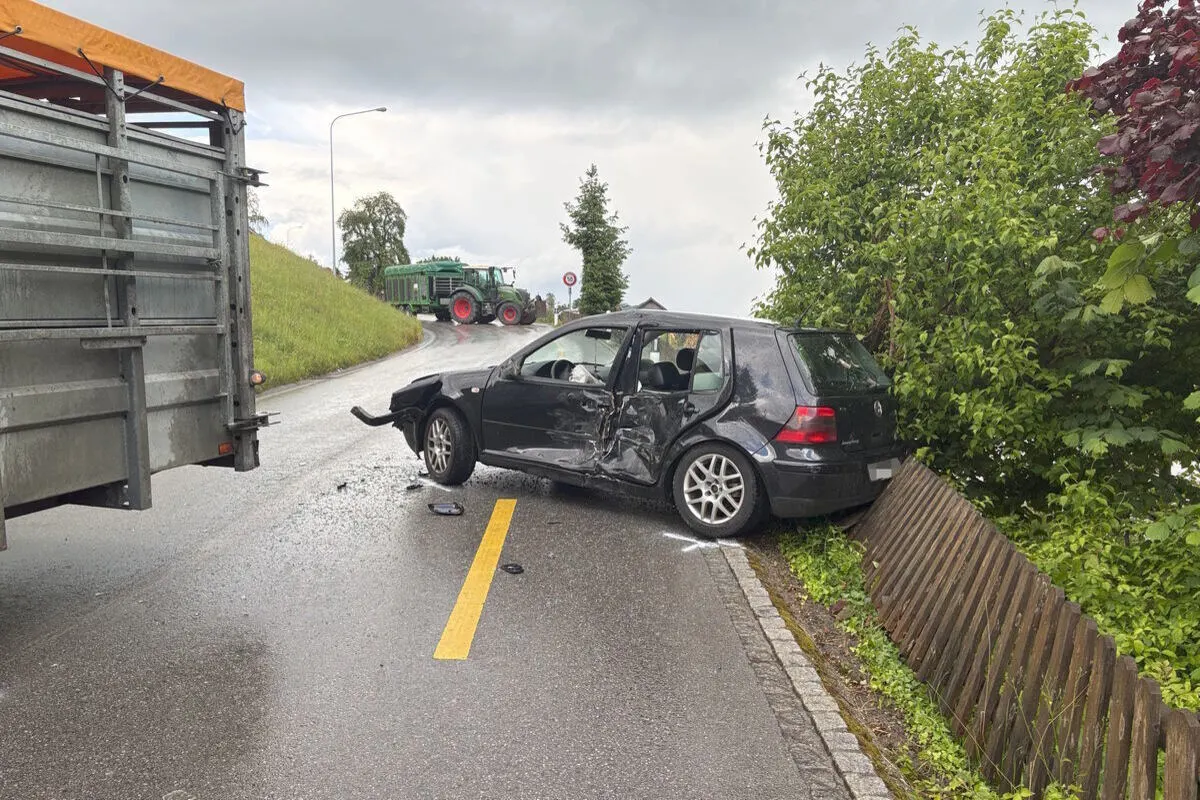 Ein schwarzes Auto mit deutlichen Beschädigungen steht auf einer nassen Straße an einer Böschung. Ein Traktor mit Anhänger ist in der Ferne sichtbar.