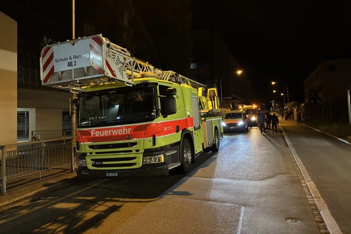 Ein Feuerwehrauto mit einer Leiter steht auf einer Straße bei Nacht. Weitere Fahrzeuge und Personen sind im Hintergrund zu sehen.