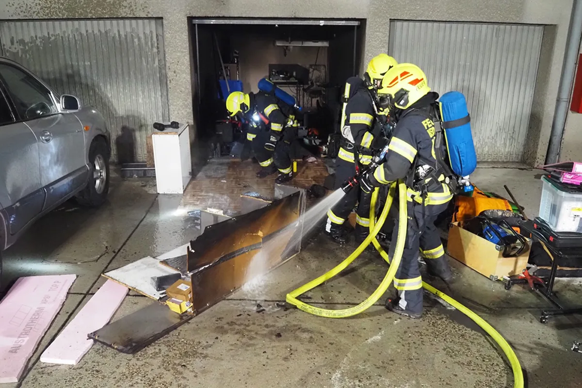 Drei Feuerwehrleute mit Atemschutzmasken löschen ein Feuer in einer Garage. Ein Auto steht daneben.
