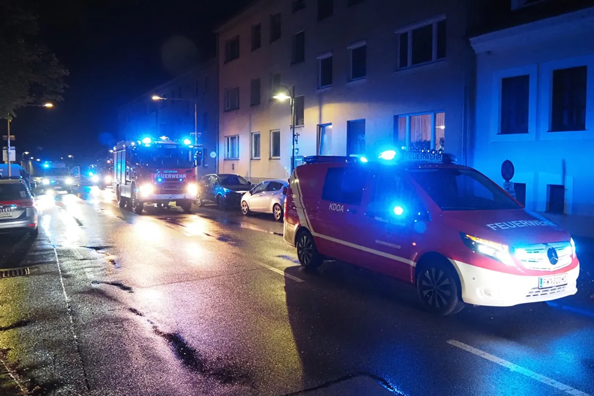 Mehrere Feuerwehrautos mit eingeschaltetem Blaulicht stehen in einer nächtlichen Wohnstraße. Die Straße ist nass und wird von Straßenlaternen beleuchtet.