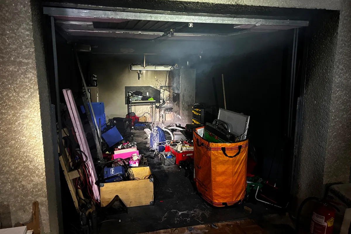 Das Bild zeigt das Innere einer Garage mit mehreren sichtbaren Brandschäden und unordentlich verstreuten Gegenständen, darunter Werkzeugkisten und ein großer orangefarbener Beutel. Ein Feuerlöscher ist am Rand erkennbar.