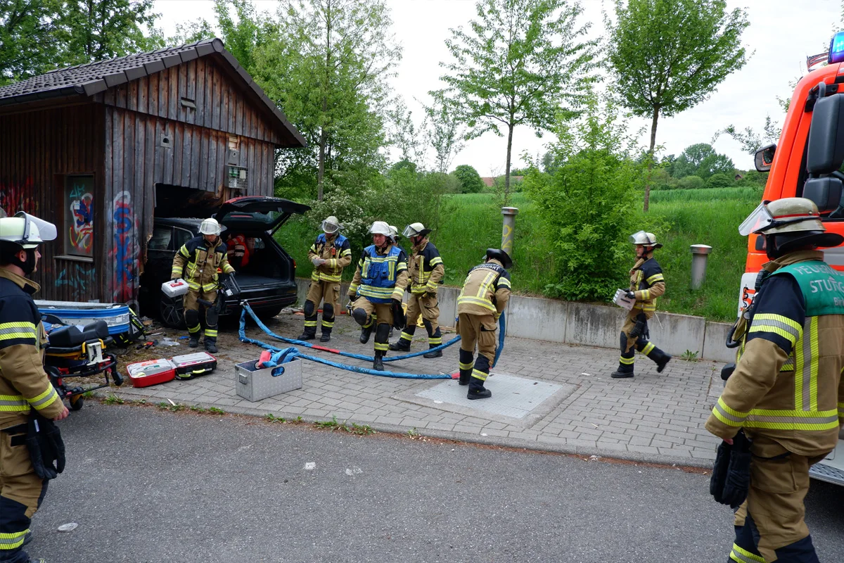 Eine Gruppe von Feuerwehrleuten arbeitet an der Unfallstelle, an der ein Fahrzeug in ein Holzgebäude gefahren ist. Ausrüstung und ein Feuerwehrauto sind ebenfalls zu sehen.