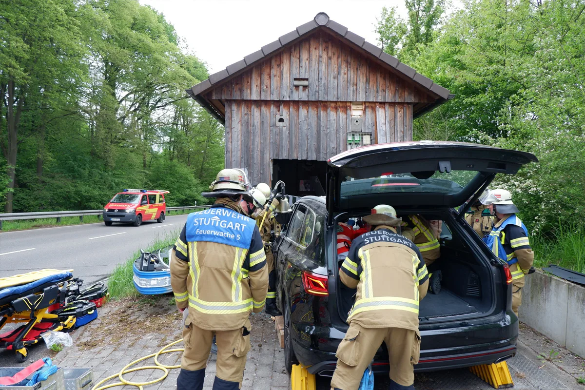Mehrere Feuerwehrleute der Feuerwehr Stuttgart arbeiten an einem schwarzen Auto, das vor einem hölzernen Gebäude steht. Einsatzfahrzeuge und Ausrüstung sind sichtbar.