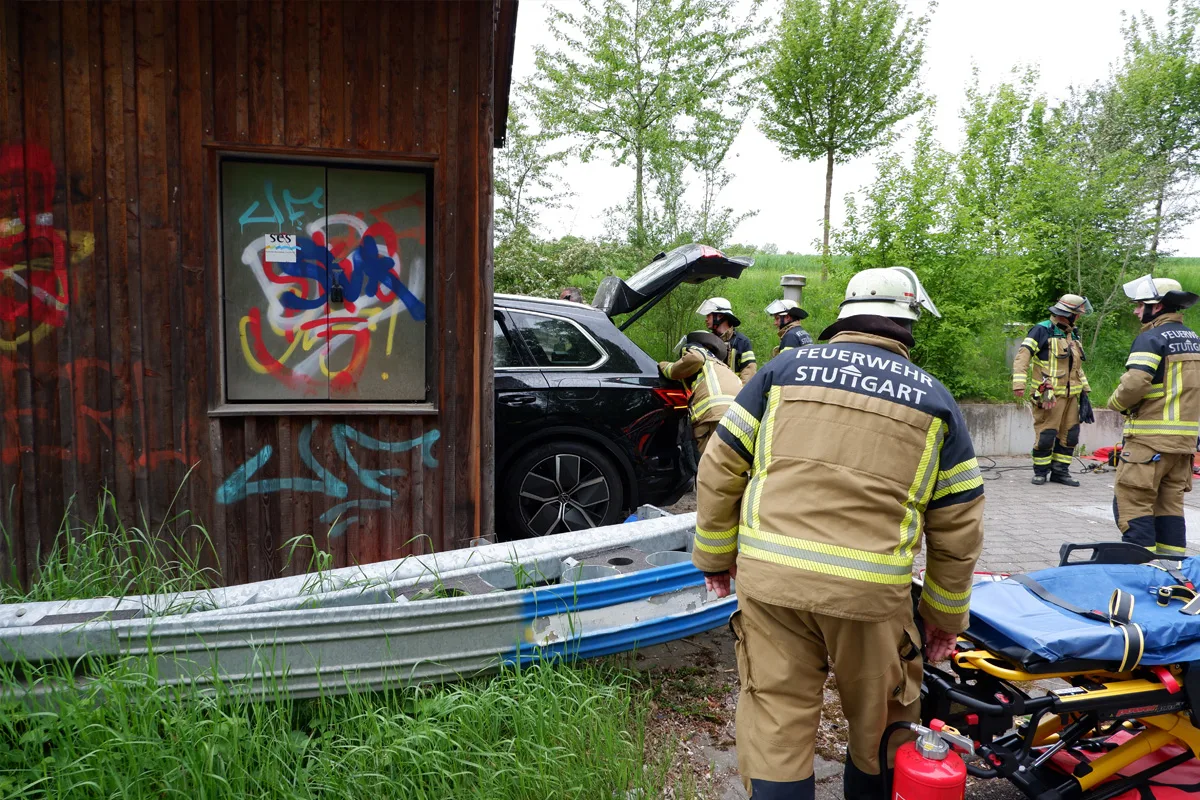 Mehrere Feuerwehrleute arbeiten an einem schwarzen Auto, das neben einer Holzwand mit Graffiti steht. Ein Tragewagen und andere Ausrüstungen sind neben ihnen.