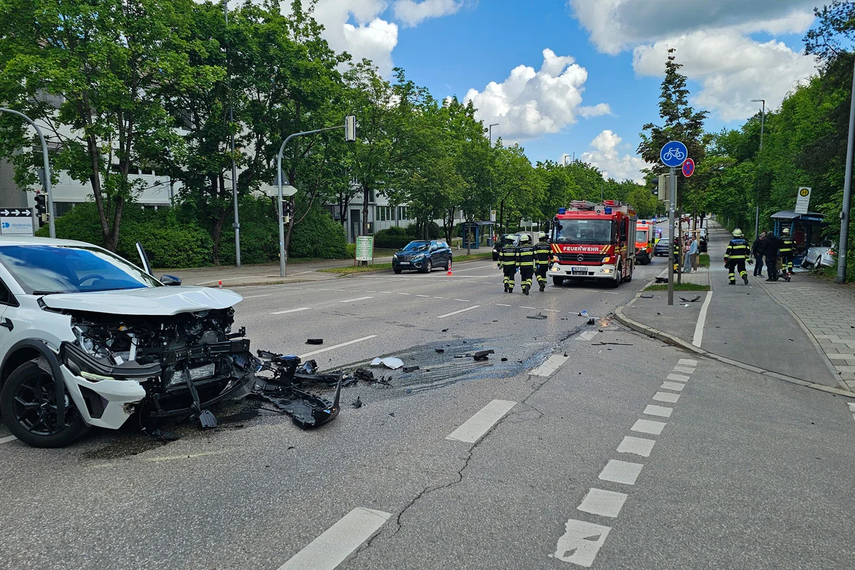 Ein stark beschädigtes weißes Auto steht links auf einer Stadtstraße. Rechts sind Feuerwehrleute und ein Feuerwehrfahrzeug zu sehen.