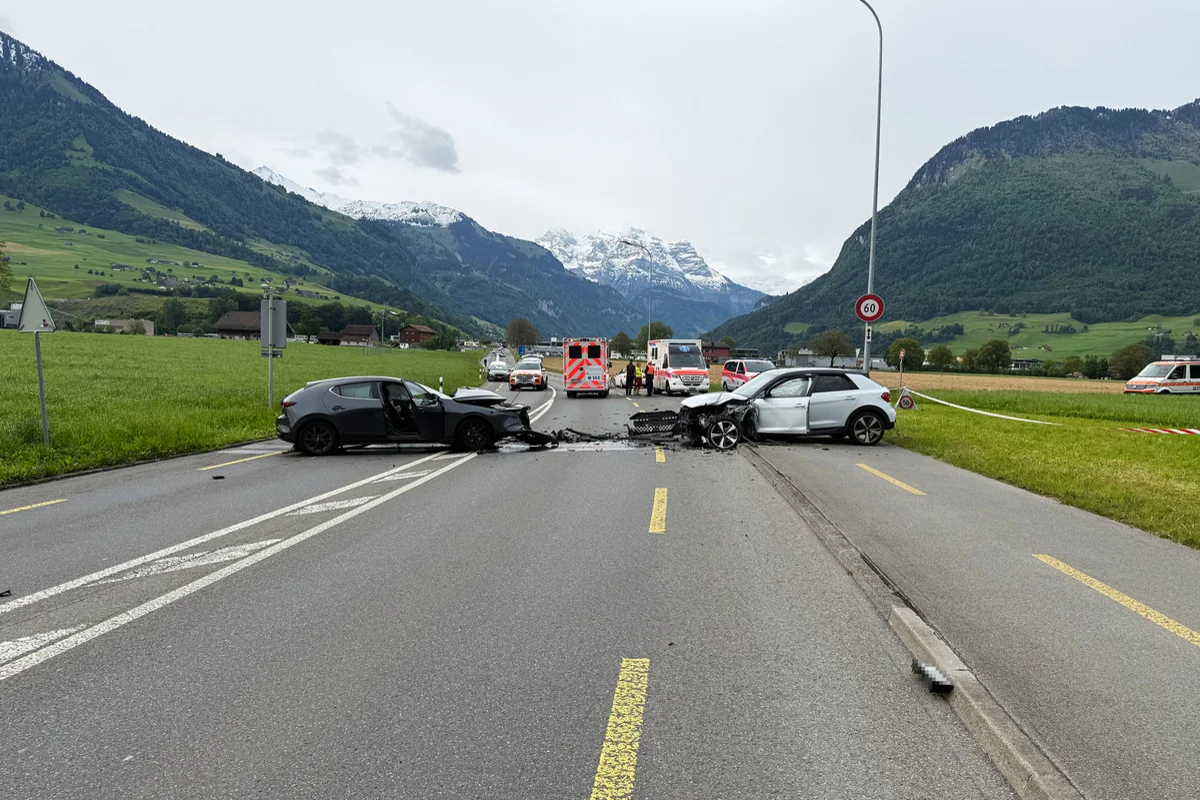 Zwei Autos sind frontal auf einer Landstraße kollidiert, Trümmer liegen verstreut. Einsatzfahrzeuge und Berge im Hintergrund sichtbar.