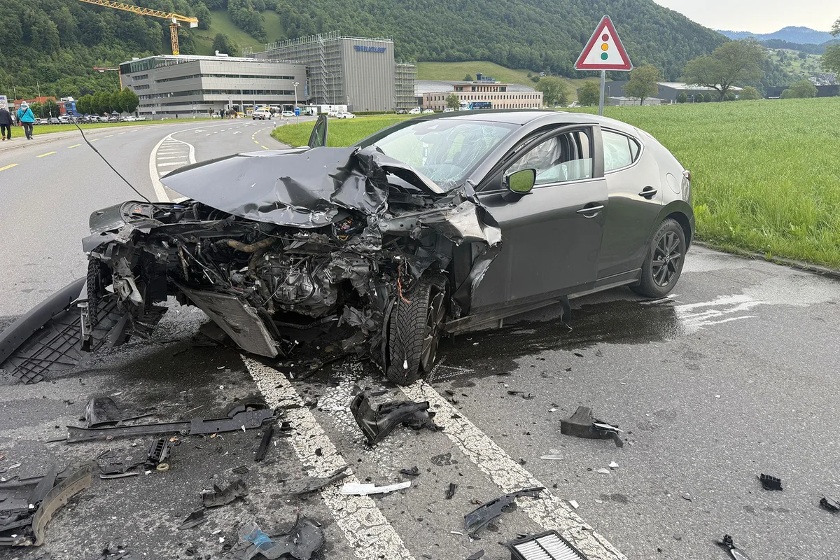 Ein Auto ist vorne stark beschädigt und steht auf einer Straße, umgeben von Trümmern. Im Hintergrund sind Grünflächen und ein Industriegebäude zu sehen.