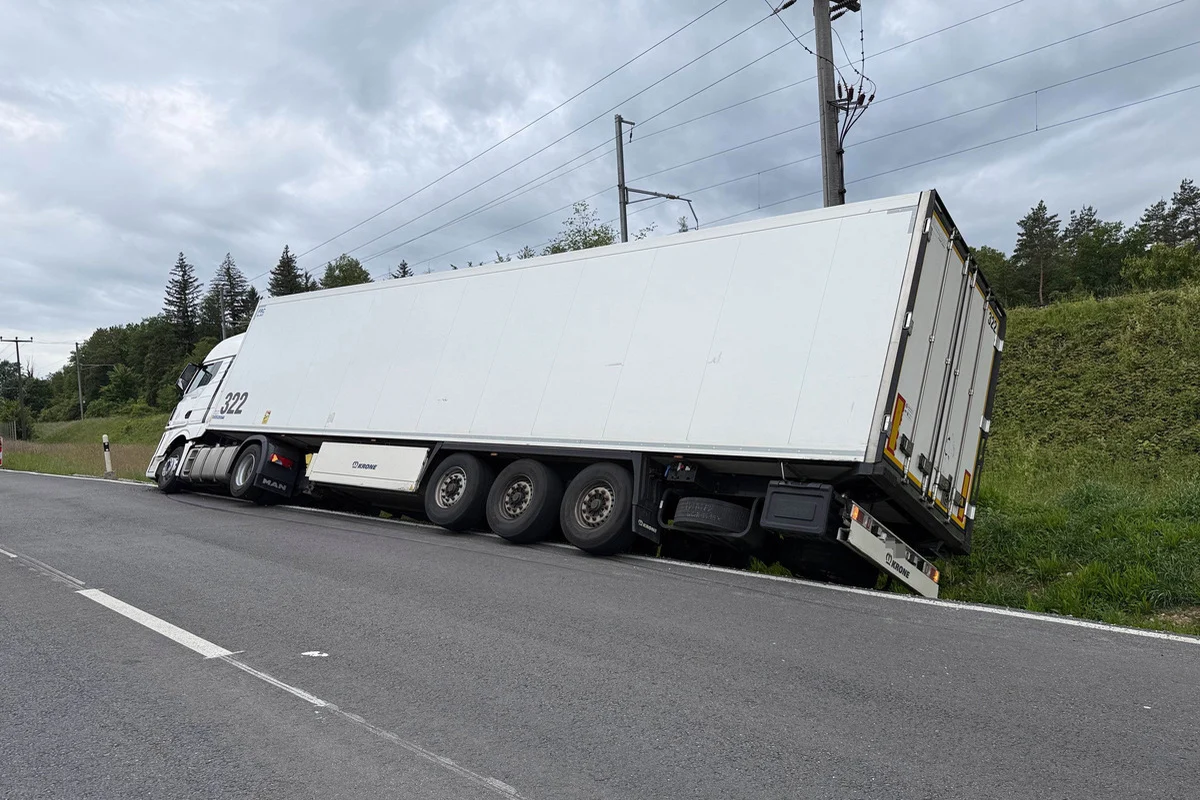 Ein schlichter weißer Lastwagen ist von der Straße abgekommen und mit der rechten Seite in eine Böschung geraten. Die Umgebung zeigt eine Landstraße mit Gras und Bäumen, während im Himmel Wolken sichtbar sind.