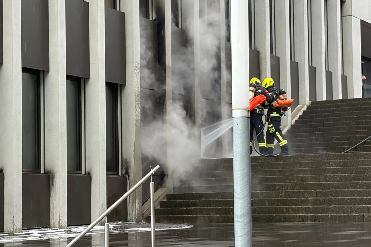 Zwei Feuerwehrleute in voller Schutzkleidung sprühen Wasser auf eine rauchende Stelle an einer Gebäudefront mit Treppen.