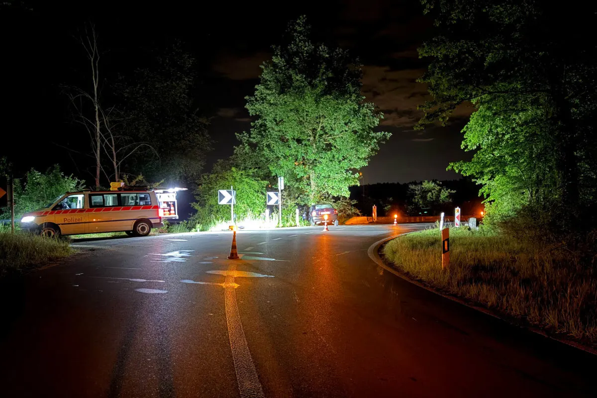 Ein Polizeifahrzeug mit eingeschaltetem Licht steht an einer kurvenreichen Kreuzung nachts. Ein weiteres Auto ist im Hintergrund neben der Straße zu erkennen.