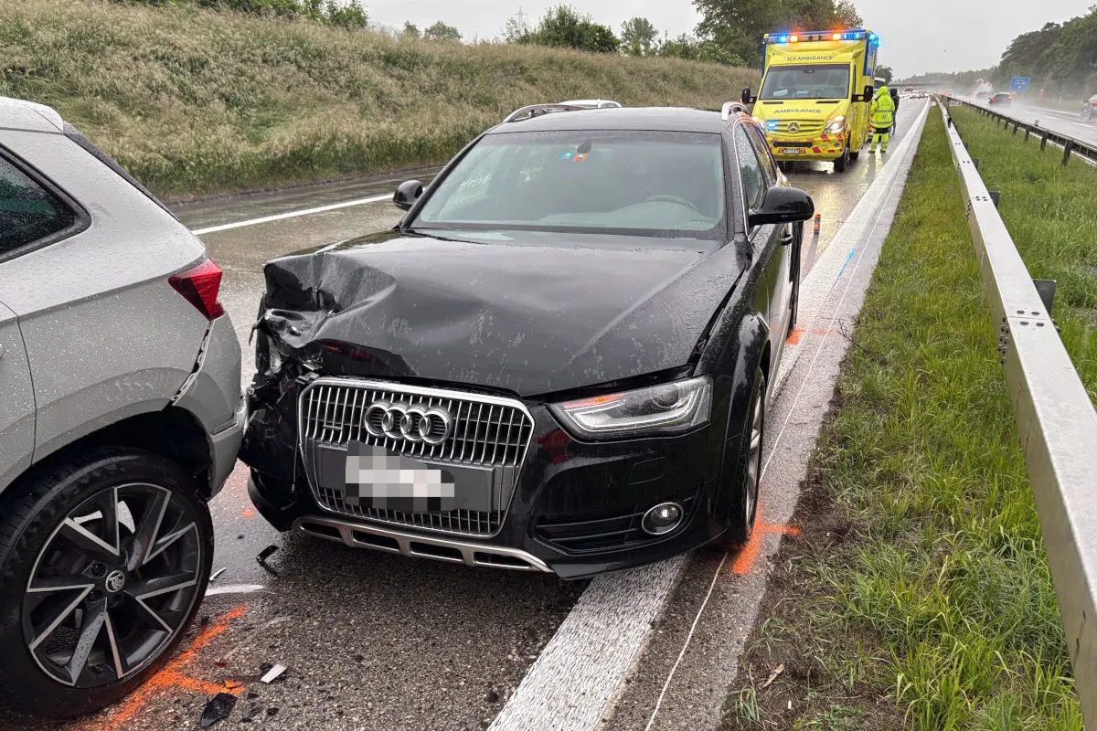 Ein schwarzes Audi mit Frontschaden steht auf dem Seitenstreifen einer Autobahn und ist auf ein graues Auto aufgefahren. Im Hintergrund ist ein Rettungswagen zu sehen.