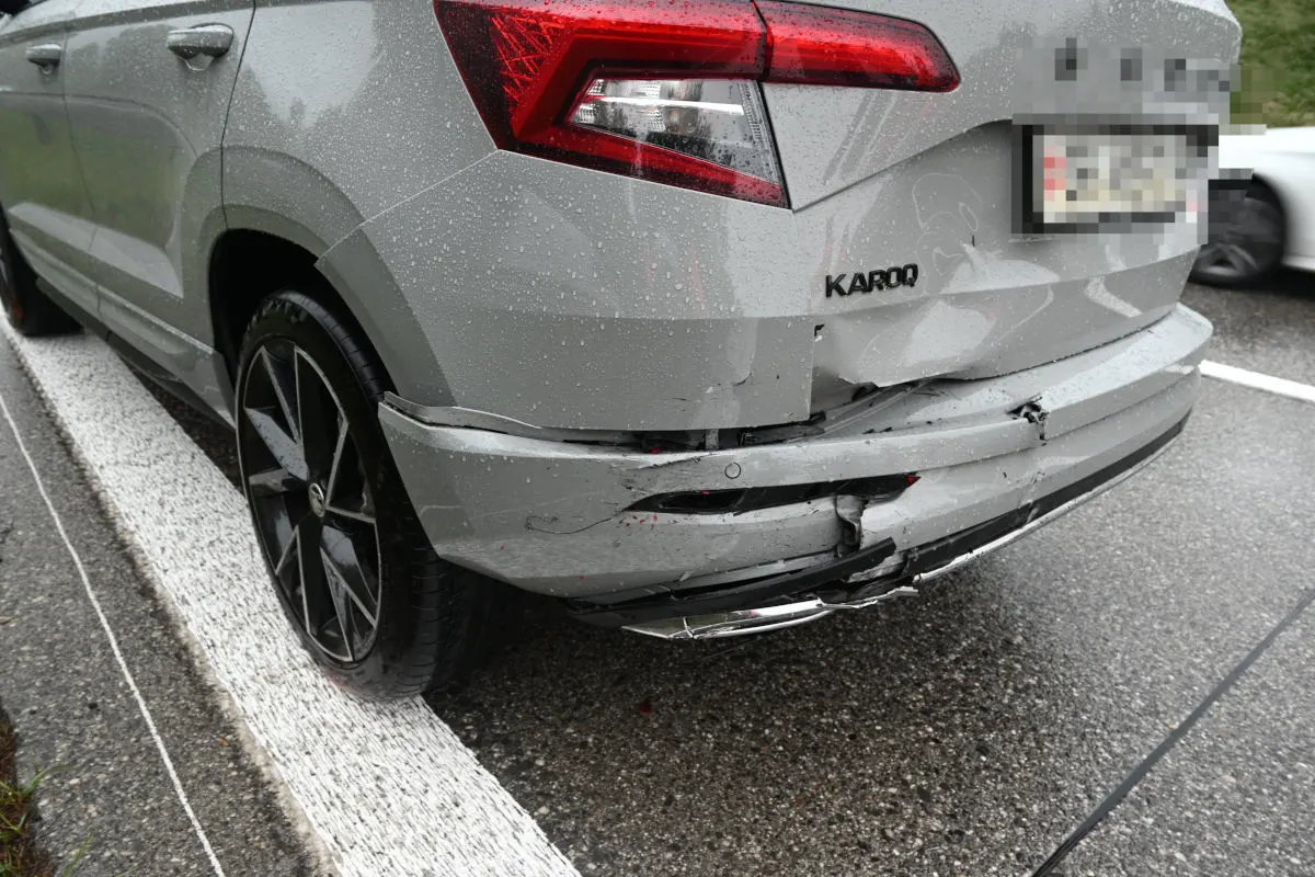 Das Bild zeigt das Heck eines grauen Autos mit deutlichen Unfallspuren am Stoßfänger. Es ist nass, was auf regnerisches Wetter hindeutet, und das Auto steht auf der Autobahn.