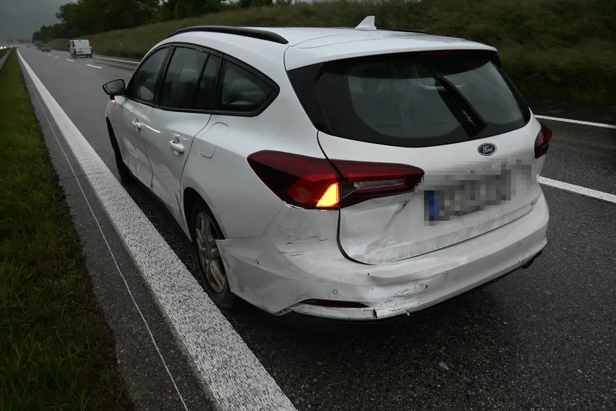 Ein weißer Ford mit deutlichen Heckbeschädigungen steht auf dem Seitenstreifen einer Autobahn bei nasser Fahrbahn.