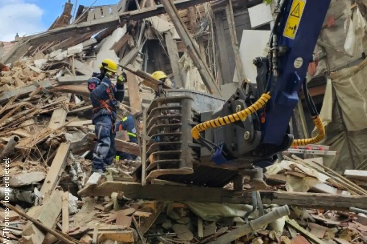 Rettungskräfte in Schutzkleidung arbeiten mit schwerem Gerät in einem Trümmerfeld eines eingestürzten Gebäudes. Holztrümmer und verbogenes Metall sind sichtbar.