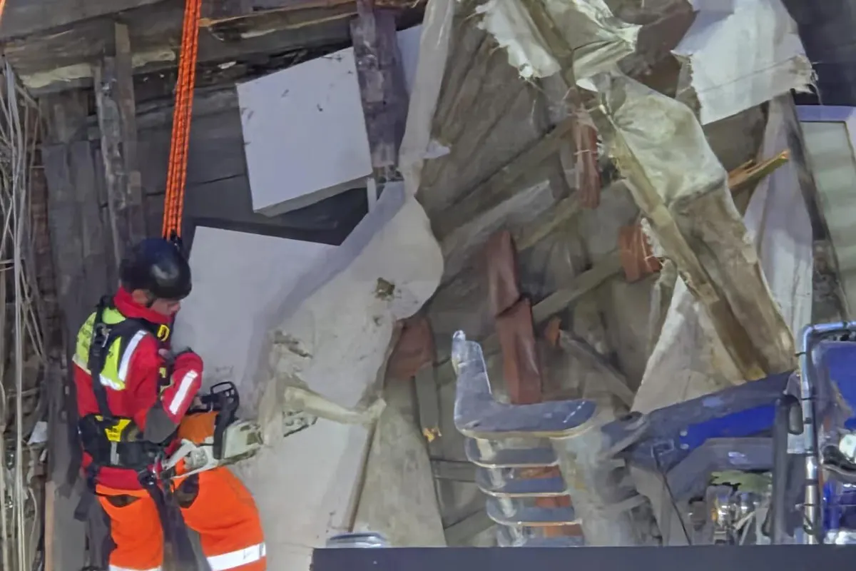 Eine Rettungskraft mit roter Schutzkleidung und Kettensäge arbeitet in einem stark beschädigten Gebäude. Herabhängende Trümmer und freiliegende Holzstrukturen umgeben die Szenerie.