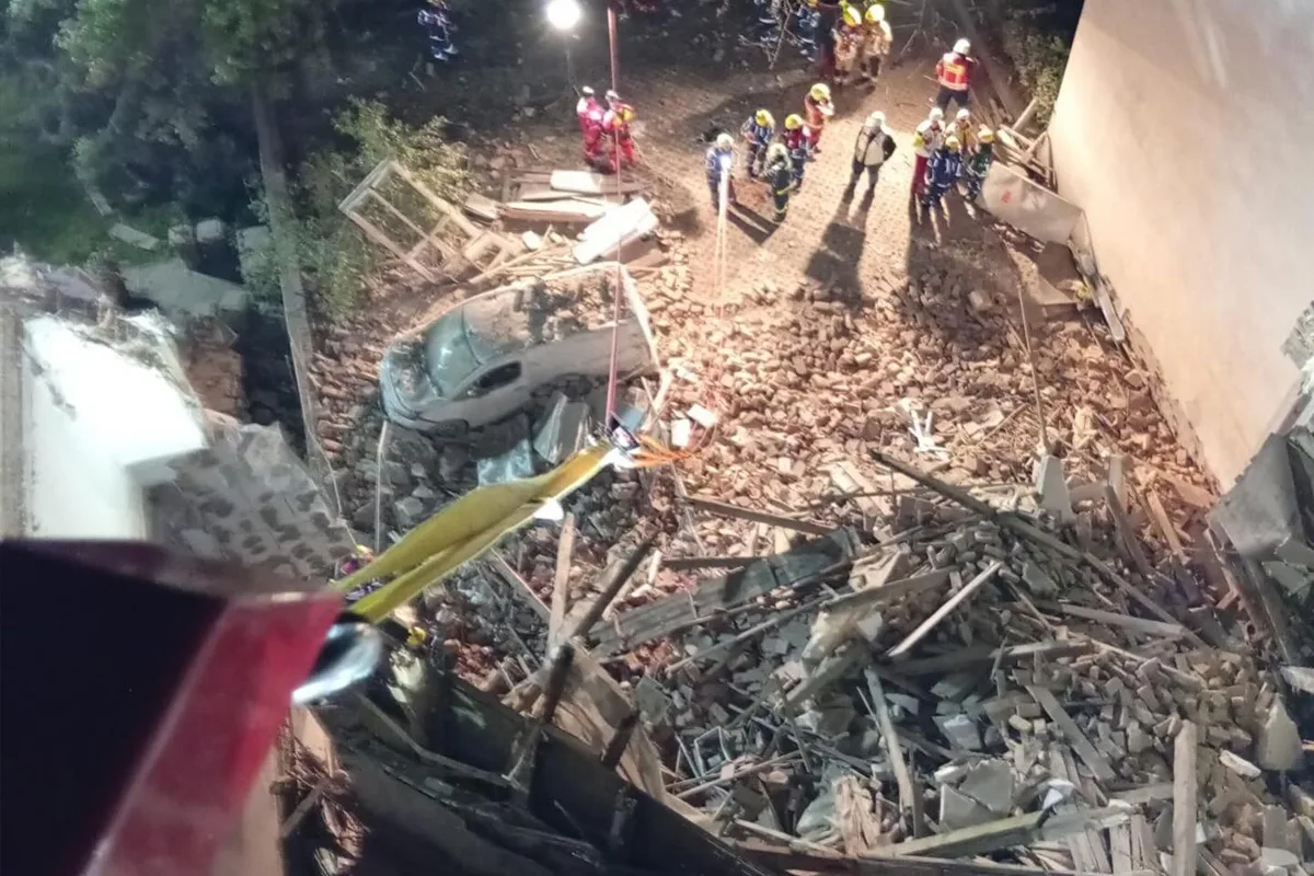 Blick von oben auf ein Fahrzeug in den Trümmern eines eingestürzten Gebäudes, umgeben von Rettungskräften bei Nacht.