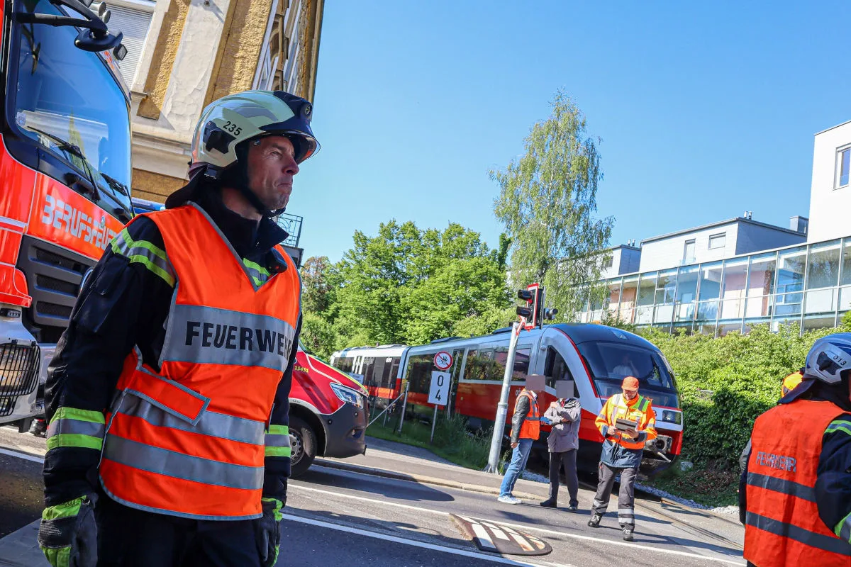 Ein Feuerwehrmann in oranger Warnweste und Helm steht neben einem roten Feuerwehrauto vor einem Zug. Weitere Einsatzkräfte sind im Hintergrund sichtbar.