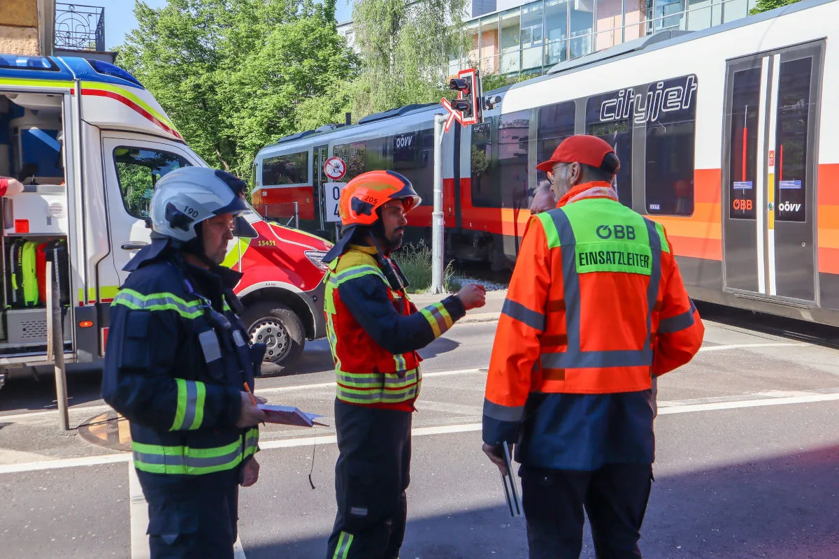 Drei Feuerwehrleute in Schutzkleidung stehen neben einem ÖBB-Zug und einem Einsatzfahrzeug, während sie miteinander sprechen und Pläne diskutieren.