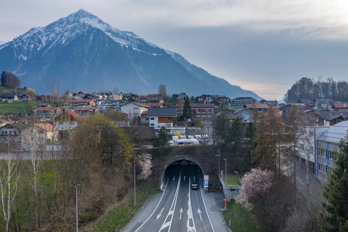 Ein Tunnel führt durch einen grünen Hügel, daneben stehen Häuser und Bäume. Im Hintergrund ragen schneebedeckte Berge empor.