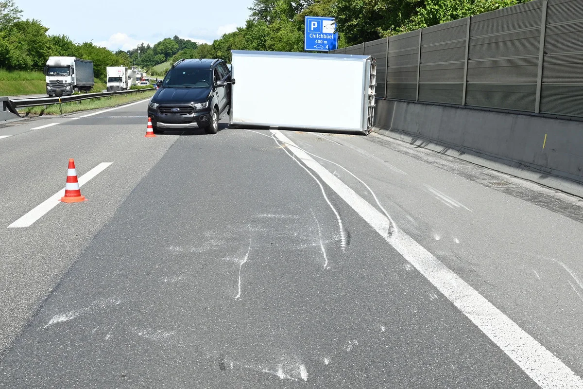 Ein großer, umgekippter Anhänger blockiert die rechte Fahrspur einer Autobahn. Ein schwarzes Fahrzeug steht daneben, mit Verkehrskegeln abgesichert.
