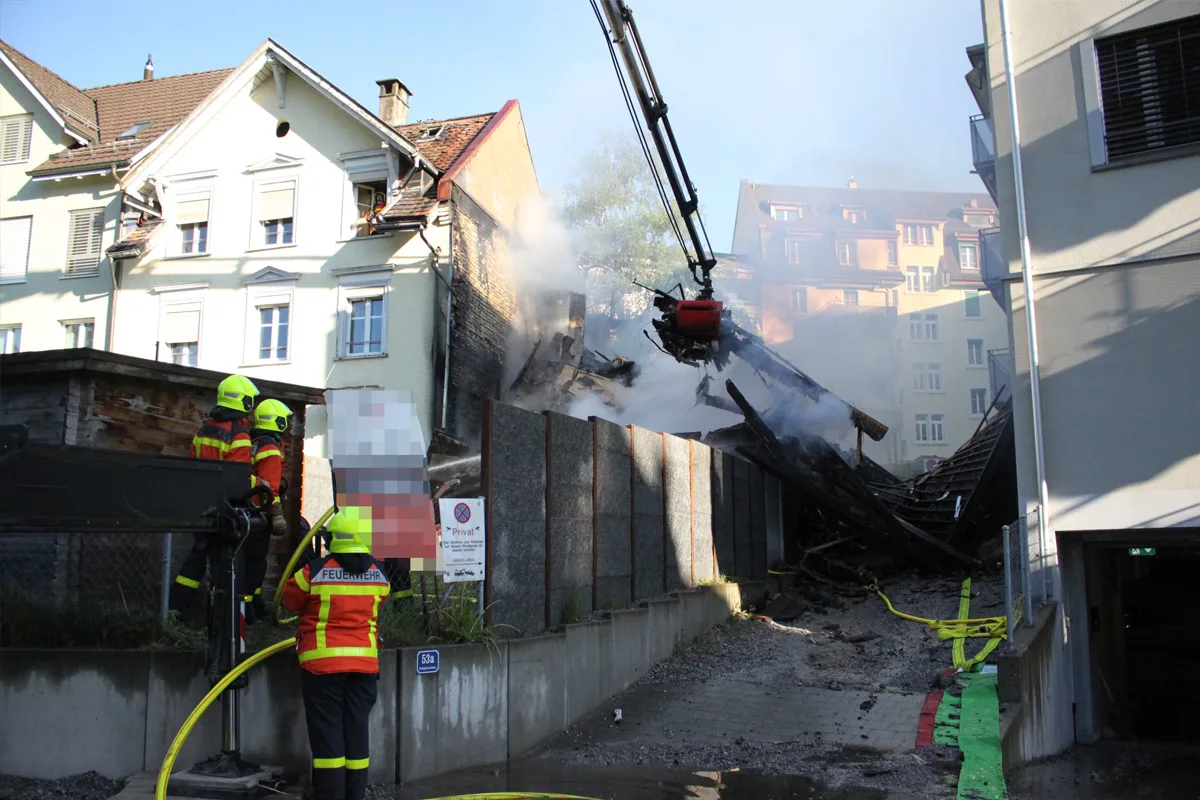 Drei Feuerwehrleute in Schutzkleidung stehen vor einem eingestürzten Gebäude, aus dem Rauch aufsteigt. Ein Kran ist im Einsatz, um die Überreste zu sichern.