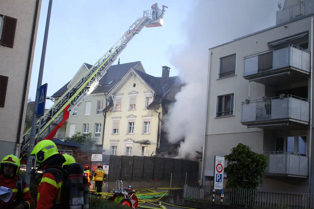 Das Bild zeigt eine Feuerwehrleiter, die zu einem Mehrfamilienhaus führt, aus dem dichter Rauch aufsteigt. Feuerwehrleute sind im Einsatz zu sehen.