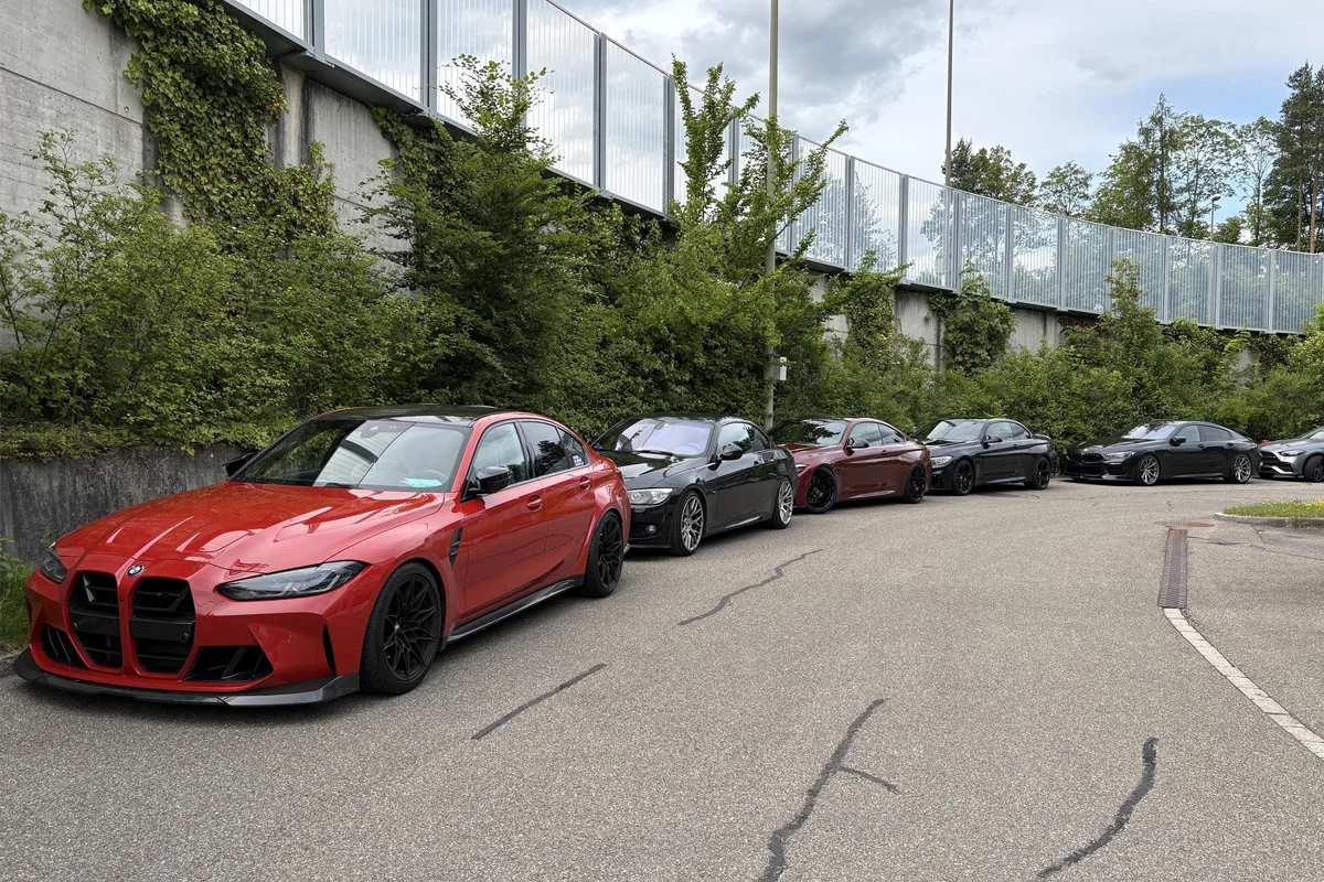 Eine Reihe von Fahrzeugen steht geparkt an einer Straße neben einer Lärmschutzwand, darunter ein auffälliger roter BMW im Vordergrund. Die Umgebung ist von grüner Vegetation gesäumt.