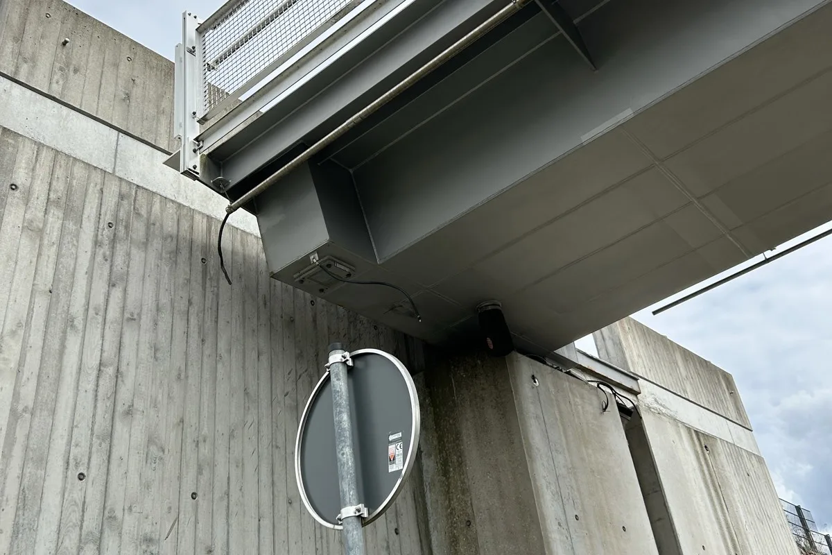 Das Bild zeigt die Unterseite einer Brücke mit Betonwand und einem runden Straßenschild. Kabel und technische Komponenten sind sichtbar.