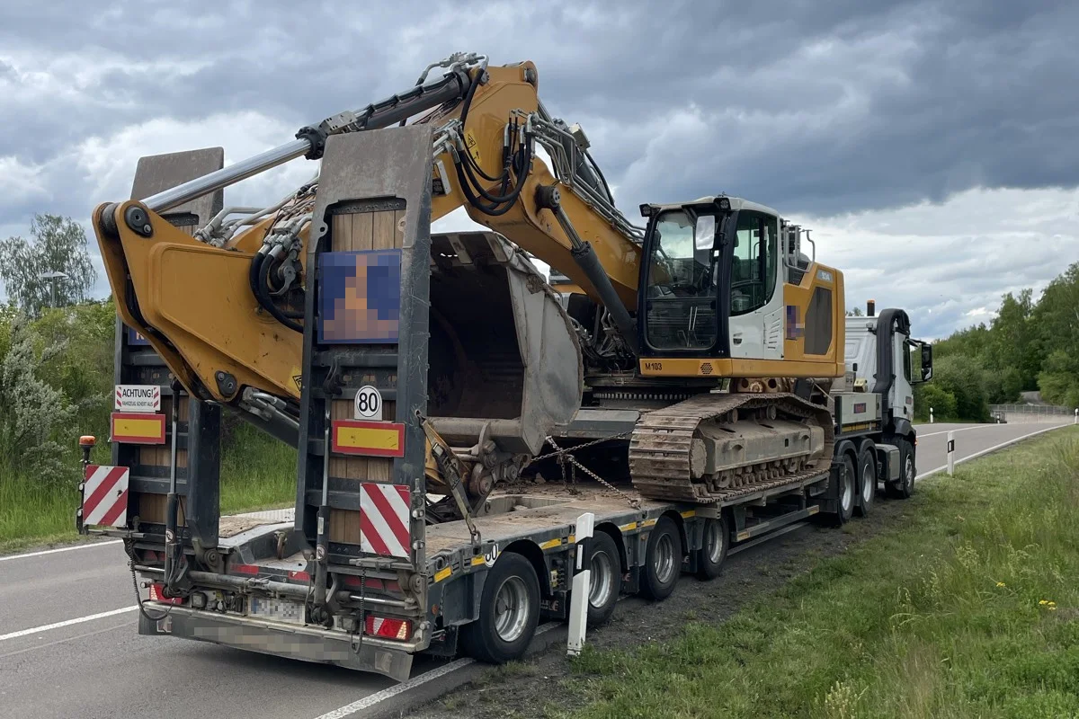 Ein großer gelb-weißer Bagger steht auf einem Sattelschlepper, der am Straßenrand unter einem bewölkten Himmel geparkt ist. Der Transporter befindet sich auf einer zweispurigen Landstraße, umgeben von Grünflächen und Bäumen.