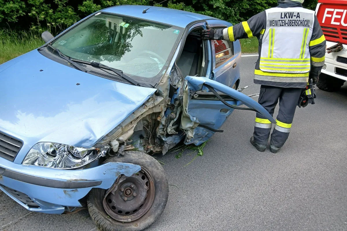 Ein stark beschädigtes blaues Auto nach einem Unfall mit einem Feuerwehrmann, der daneben steht. Der Vorderreifen ist zerstört und die Karosserie stark eingedrückt.