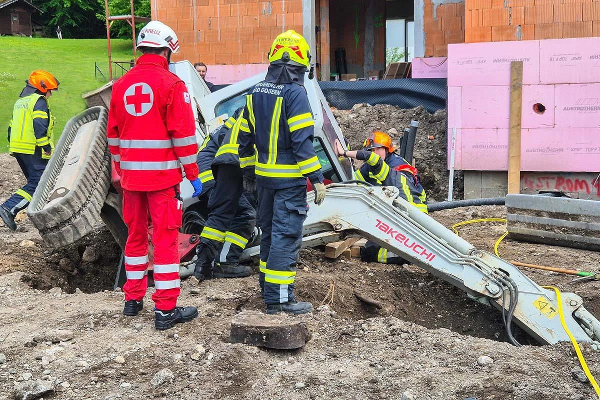 Ein Team von Feuerwehrleuten und einem Mitglied des Rettungsdienstes arbeiten gemeinsam an einem Unfallort auf einer Baustelle, bei dem ein Bagger involviert ist.