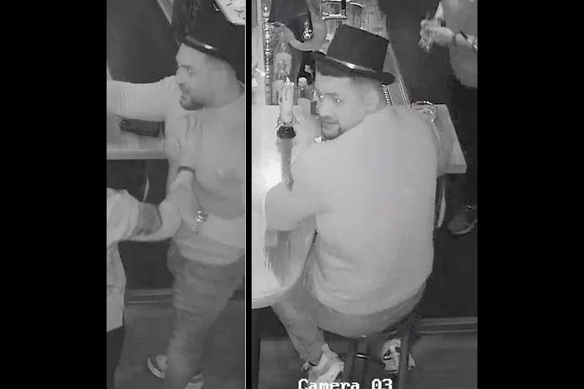Ein Mann mit einem Zylinder sitzt an der Theke in einer Bar und schaut in Richtung der überwachenden Kamera. Mehrere Personen sind teilweise sichtbar.