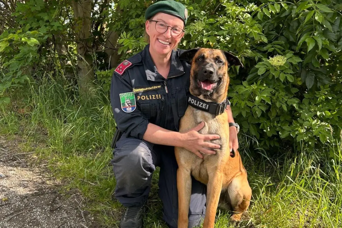 Eine weibliche Polizistin, die eine dunkelblaue Uniform trägt, kniet neben ihrem Diensthund auf einem Grünstreifen. Beide wirken entspannt und vertraut.