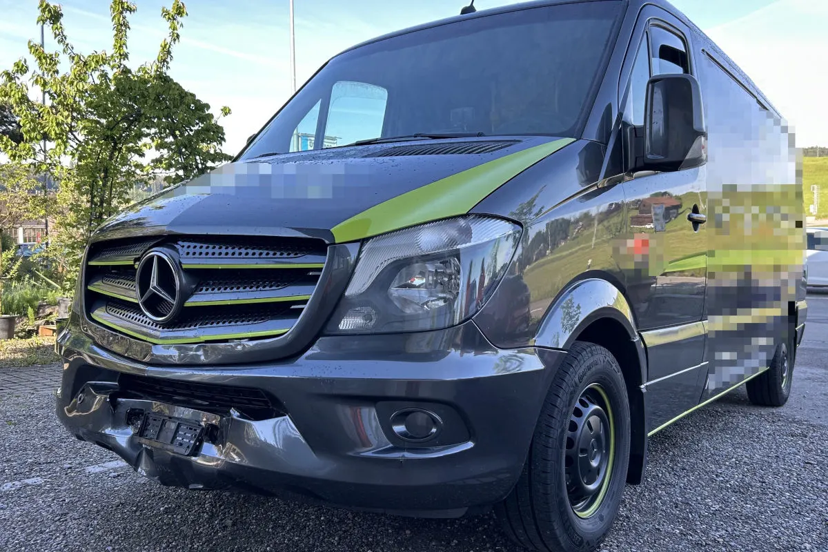 Ein silberner Mercedes-Transporter mit grünen Akzenten zeigt deutliche Dellen und Kratzer an der Frontstoßstange. Im Hintergrund sind Bäume und ein Gebäude sichtbar.
