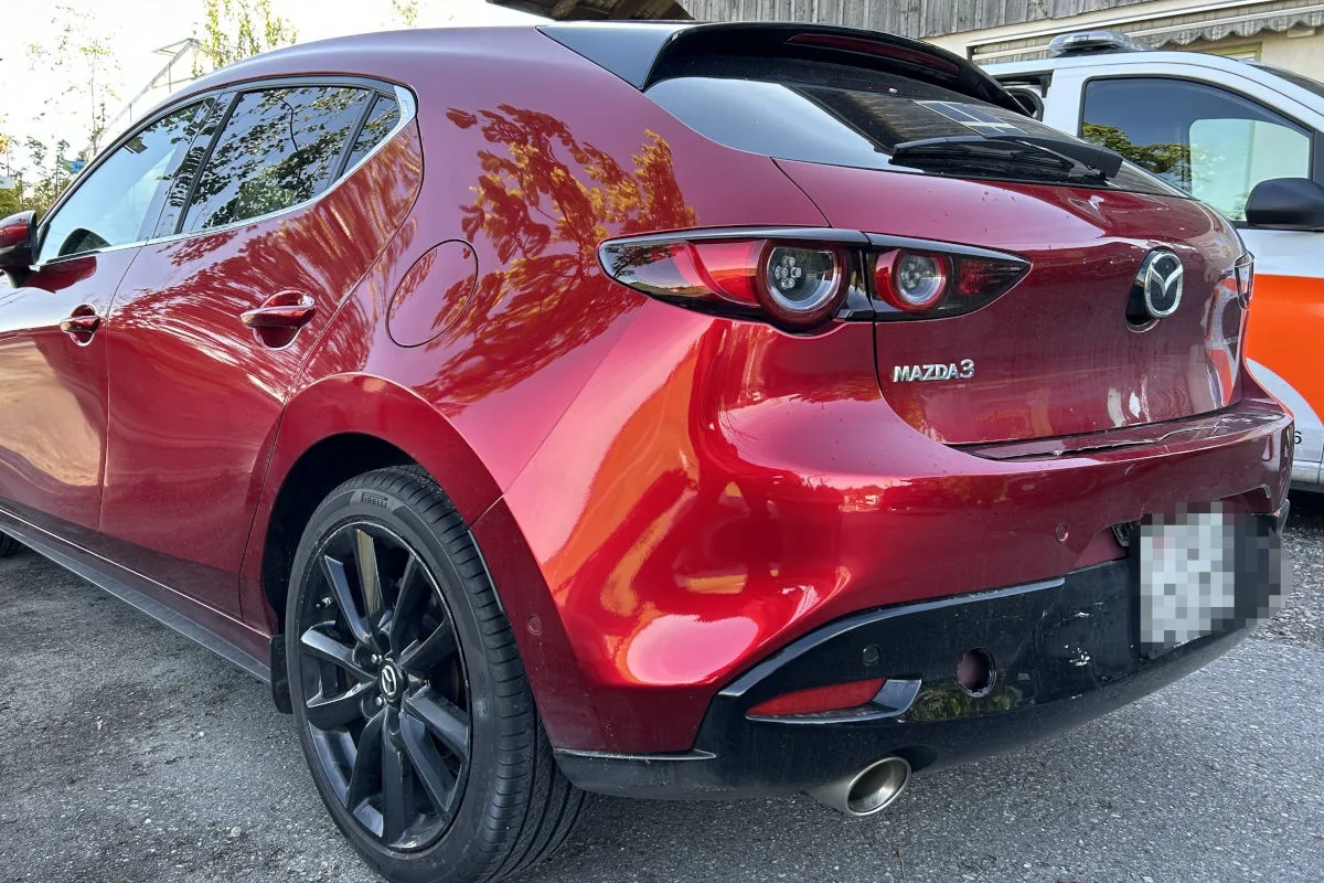 Ein roter Mazda3 parkt mit sichtbarem Schaden am Heck in einem urbanen Gebiet neben einem anderen Fahrzeug.