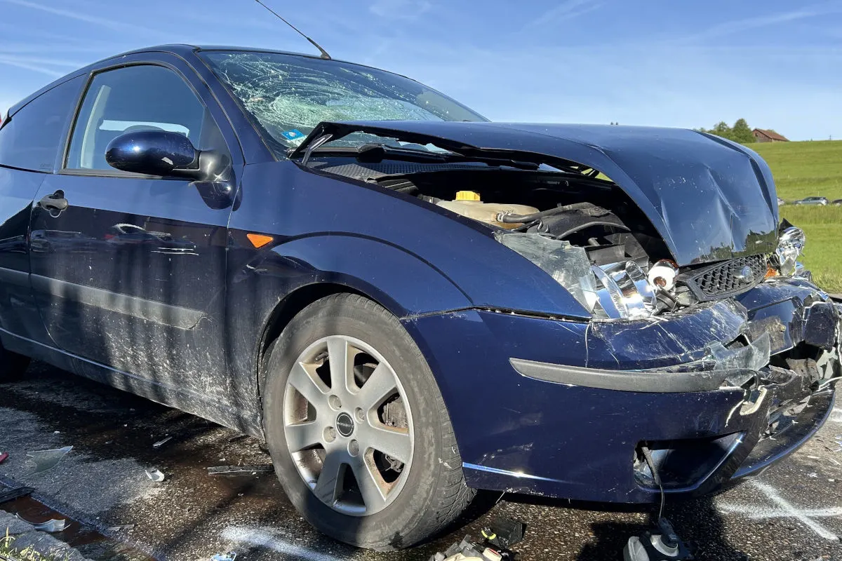 Ein blaues Auto mit schwer beschädigter Front steht auf einer Landstraße. Die Sonne scheint und der Himmel ist klar.
