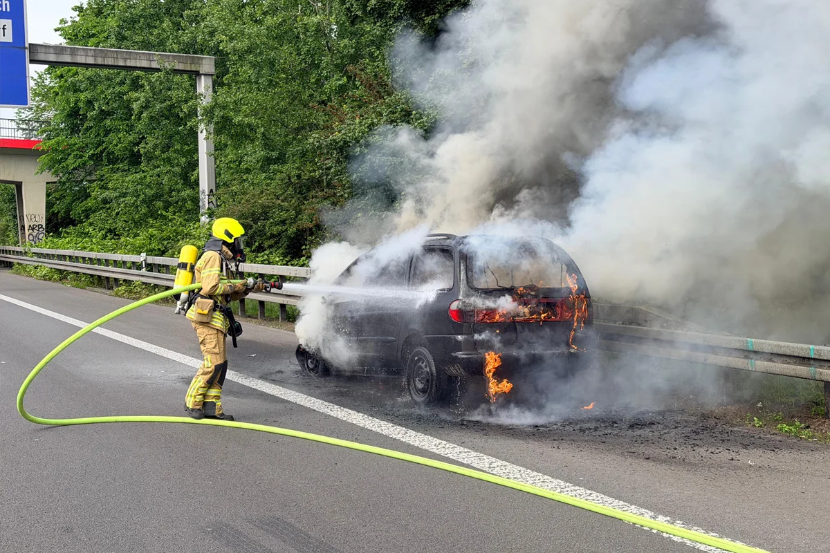 Ein Feuerwehrmann in voller Ausrüstung löscht ein Auto, das auf einer Autobahn brennt. Dicker Rauch steigt auf.