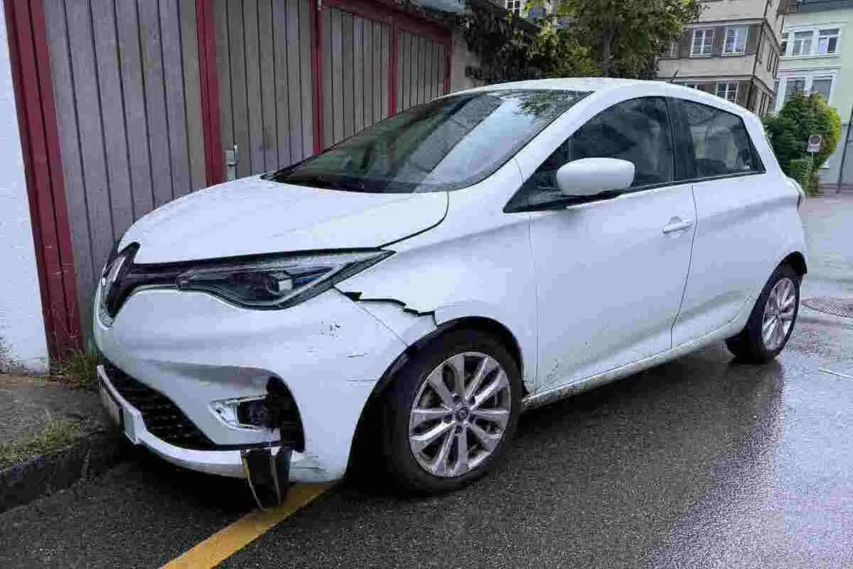 Ein weißes Auto mit sichtbarem Schaden an der vorderen Stoßstange und am Kotflügel steht am Straßenrand.