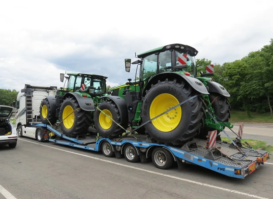 Zwei grüne Traktoren werden auf einem blauen Anhänger transportiert. Ein Fahrzeug steht offen daneben.