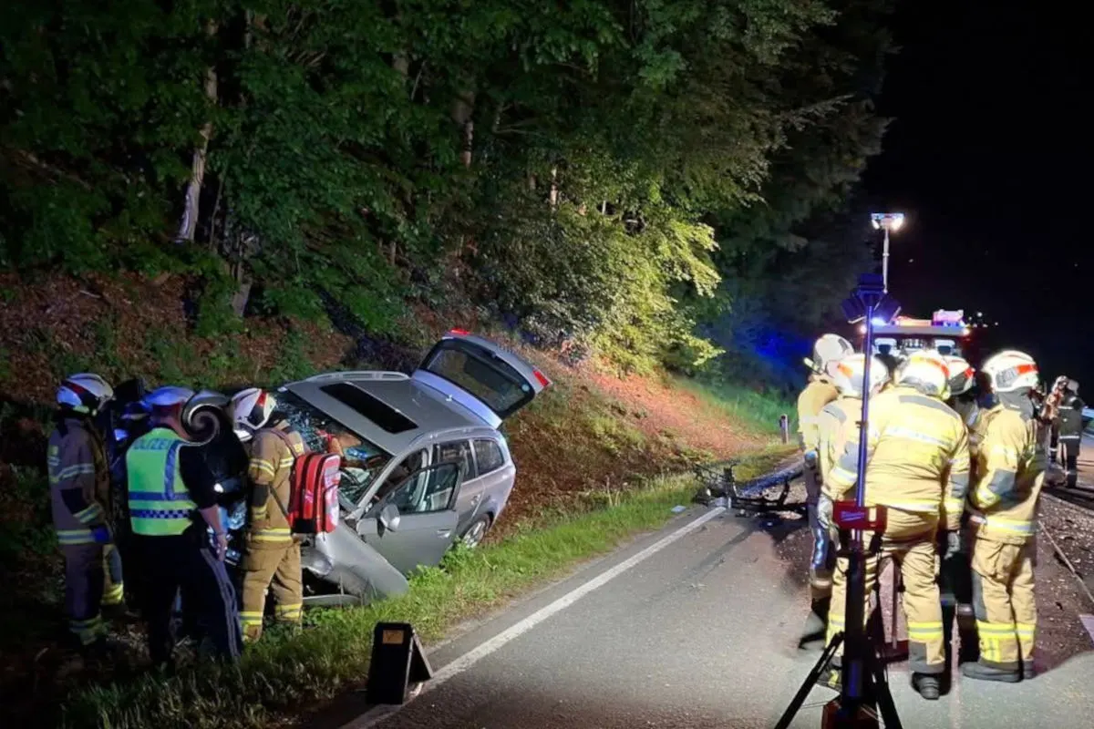 Bei Nacht stehen mehrere Feuerwehrleute an einem grauen Auto, das von der Straße abgekommen und im Graben gelandet ist. Sie arbeiten an der Unfallstelle unter einer Straßenlaterne und das Auto hat sichtbare Beschädigungen auf der Beifahrerseite.