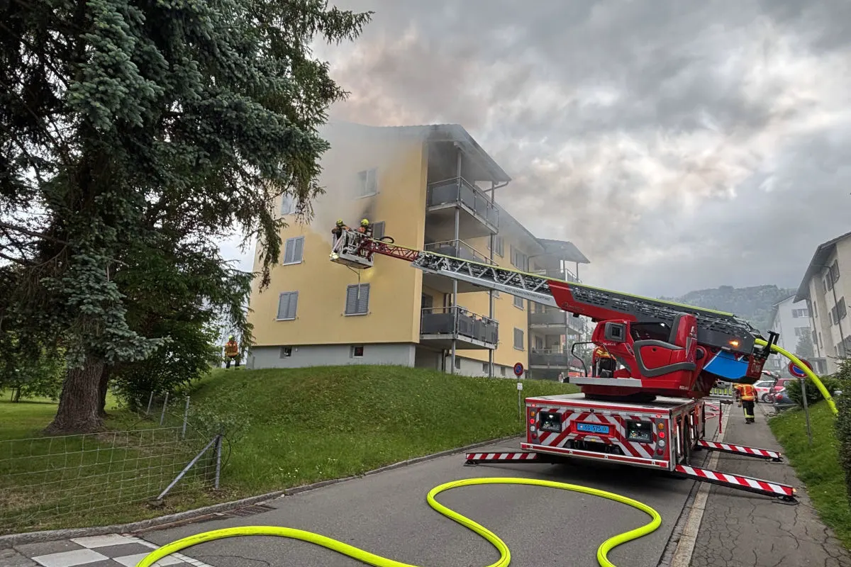 Ein großes gelbes Wohnhaus brennt; Feuerwehrleute sind auf einer Leiter und löschen den Brand mit Schläuchen.