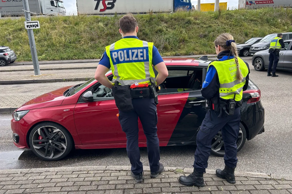 Zwei Polizisten in neongelben Westen stehen bei einem roten Auto auf einer Autobahn. Im Hintergrund sind weitere Polizeifahrzeuge zu sehen.