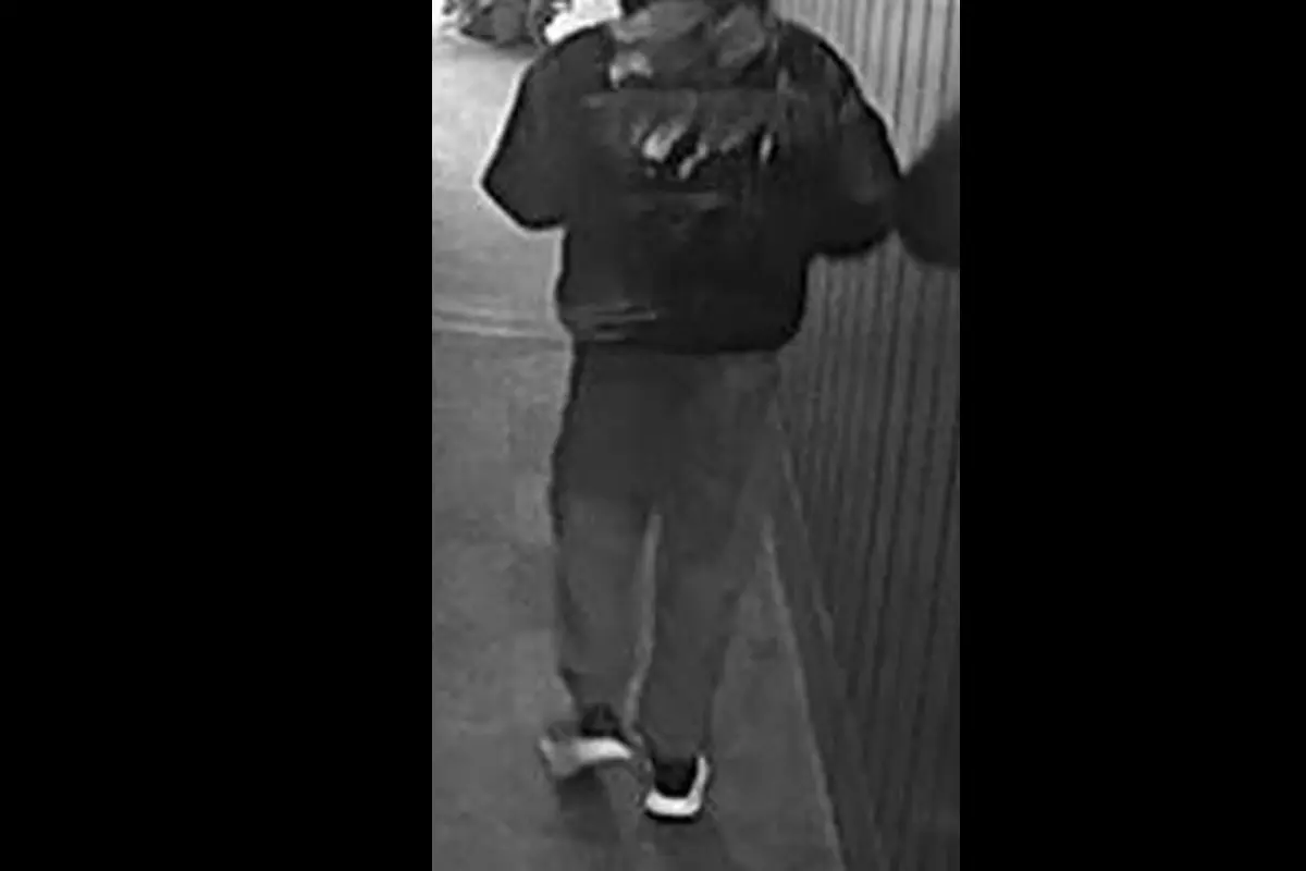 Die Aufnahme zeigt eine Person von hinten, die in einem Flur spazieren geht, bekleidet mit einer dunklen Jacke und heller Hose. Die Umgebung scheint überwacht zu sein.